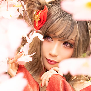 満開の桜に囲まれ、咲いた花を一房手にした赤い着物の女性の写真。